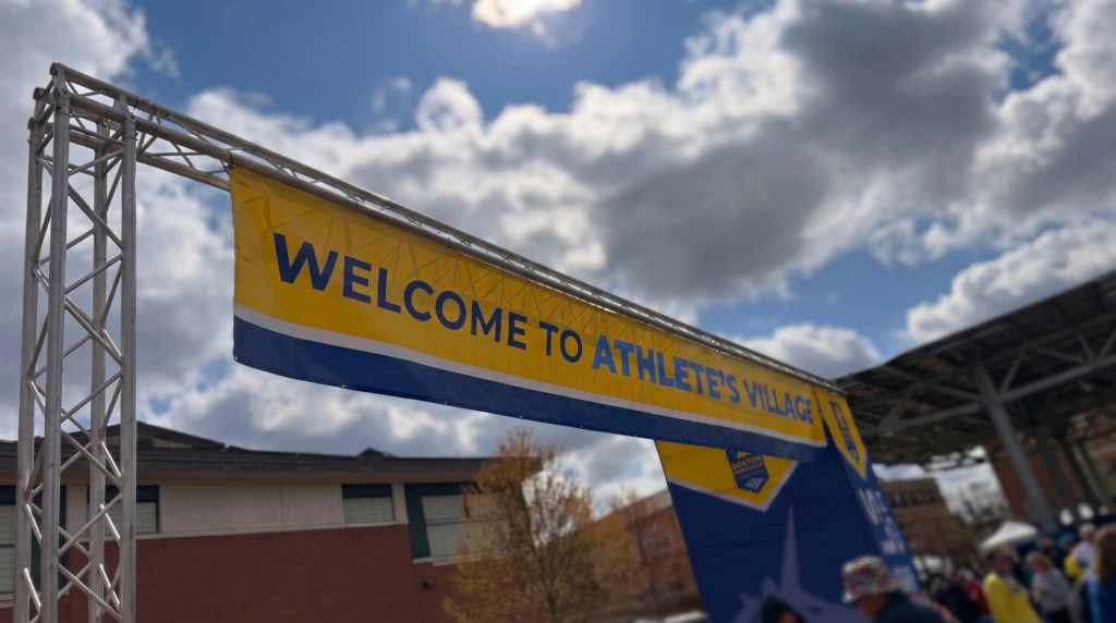 Banner reading “Welcome to Athlete’s Village” in Hopkinton under a bright race-morning sky.